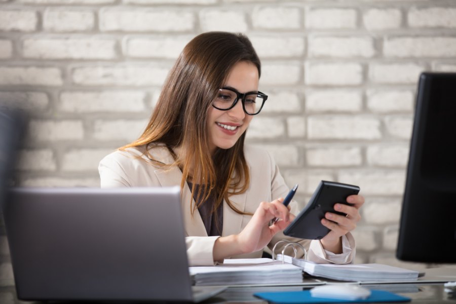 En kvinde med langt, brunt hår, iført en lys blazer og briller, smiler mens hun ser på en mobiltelefon. I baggrunden er der en bærbar computer og en stationær computer, og et par bunke papirer ligger på bordet. Væggene er dækket af lyse mursten, som giver en moderne kontorstemning.